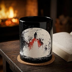 Peut inclure: Un mug en céramique noire orné d'un motif représentant une navette spatiale décollant devant une grande lune blanche texturée. Le design présente une flamme rouge vive à la base de la navette, contrastant avec le fond sombre du mug.
