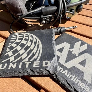 May include: Two black slate coasters with white engraved logos. One coaster features the United Airlines logo and the word "UNITED". The other coaster displays the AA logo and the words "American Airlines". A headset and audio jack are also visible.