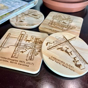 May include: Four wooden coasters with engraved airport diagrams. The coasters feature the airports of Minneapolis-St. Paul, Chicago O'Hare, Charlotte/Douglas Intl, and LaGuardia in New York. The coasters are made of light-colored wood.