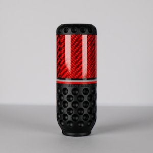 Puede incluir: Pomo de palanca de cambios Aero Design. La sección superior presenta un patrón de tejido de fibra de carbono rojo con detalles en negro. La sección media tiene una franja roja, y la sección inferior es negra con una textura con hoyuelos. El texto "AERO DESIGN" es visible.
