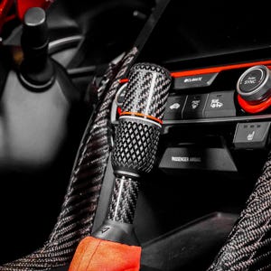 May include: A carbon fibre gear shift knob with an orange accent ring is visible inside a car. The shift boot is bright orange. The dashboard has climate control buttons and a push-to-sync dial. The interior is black.