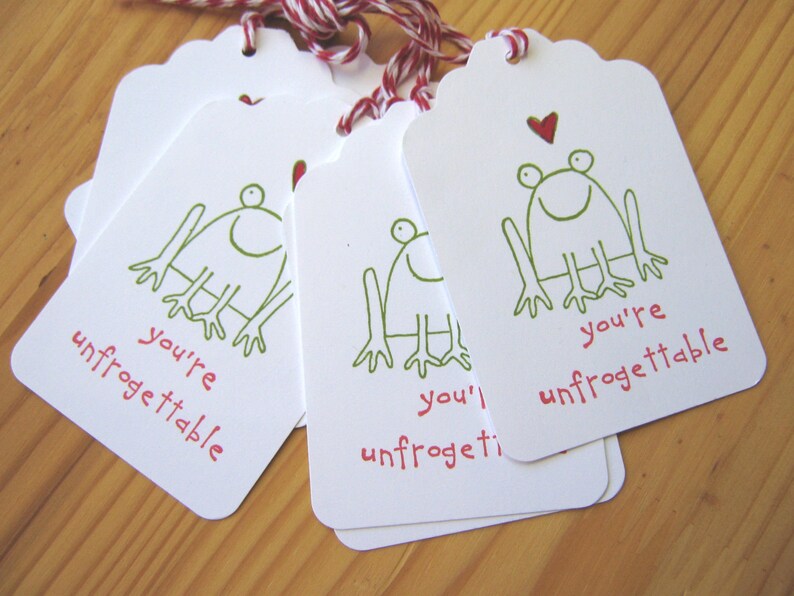 May include: Five white gift tags with a green frog illustration and the text "you're unfrogettable". The tags are tied with red and white twine.