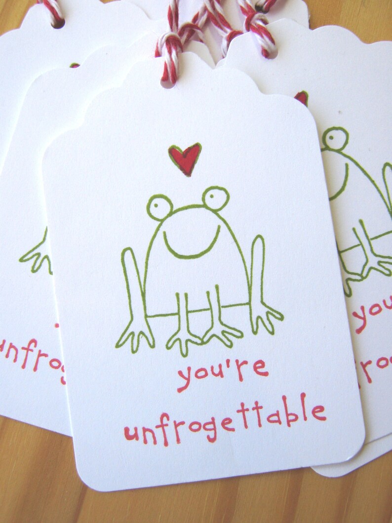 May include: A white gift tag with a green frog illustration and a red heart. The tag says "you're unfrogettable".