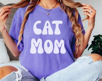 May include: A woman wearing a purple t-shirt with the text "CAT MOM" in white, retro-style lettering. She is wearing light blue jeans and has long blonde hair.