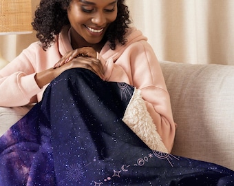 Puede incluir: Una manta azul oscuro con un diseño celestial con estrellas, lunas y soles. La manta tiene un forro suave y blanco tipo sherpa. La manta está sobre una persona sentada en un sofá beige.