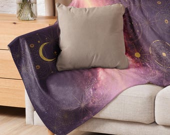 May include: A throw blanket with a celestial design featuring a purple and pink galaxy pattern, gold star and moon accents. A neutral-colored square pillow rests on the blanket. The blanket is draped over a light-colored sofa.
