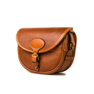 May include: A brown leather satchel bag with a buckle closure and a flap with a decorative oval. The bag has a long strap for carrying over the shoulder.