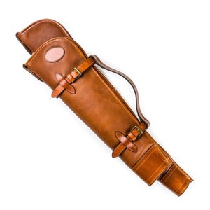 Puede incluir: Un estuche de cuero marrón para armas con asa y dos correas con hebilla. El estuche tiene una parte superior redondeada y un pequeño parche ovalado. El cuero es de color marrón medio cálido.