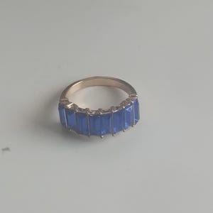 May include: A gold-toned ring featuring a row of rectangular, deep blue gemstones. The ring's band is smooth and polished, and the gemstones are set closely together, creating a continuous line of color. The ring is a piece of jewelry.