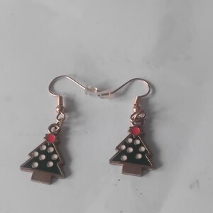 Könnte beinhalten: Ein Paar Weihnachtsbaum-Ohrringe. Jeder Ohrring zeigt einen grünen Emaille-Baum mit weißen Punktakzenten, einem roten Stern und einem braunen Stamm. Die Ohrringe haben goldfarbene Haken.