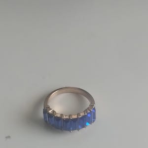 May include: A gold-toned ring featuring a row of rectangular, deep blue gemstones. The ring's band is a simple, polished metal, and the gemstones are set closely together, creating a continuous line of color. The ring is a piece of jewelry.