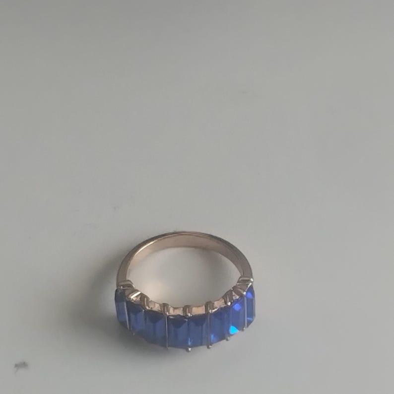 May include: A gold-toned ring featuring a row of rectangular, deep blue gemstones. The ring's band is a simple, polished metal, and the gemstones are set closely together, creating a continuous line of color. The ring is a piece of jewelry.