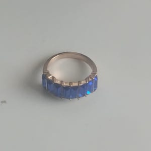 May include: A gold-toned ring featuring a row of rectangular, deep blue gemstones. The ring's band is a simple, polished metal, and the gemstones are set closely together, creating a continuous line of color. The ring is displayed on a light blue surface.