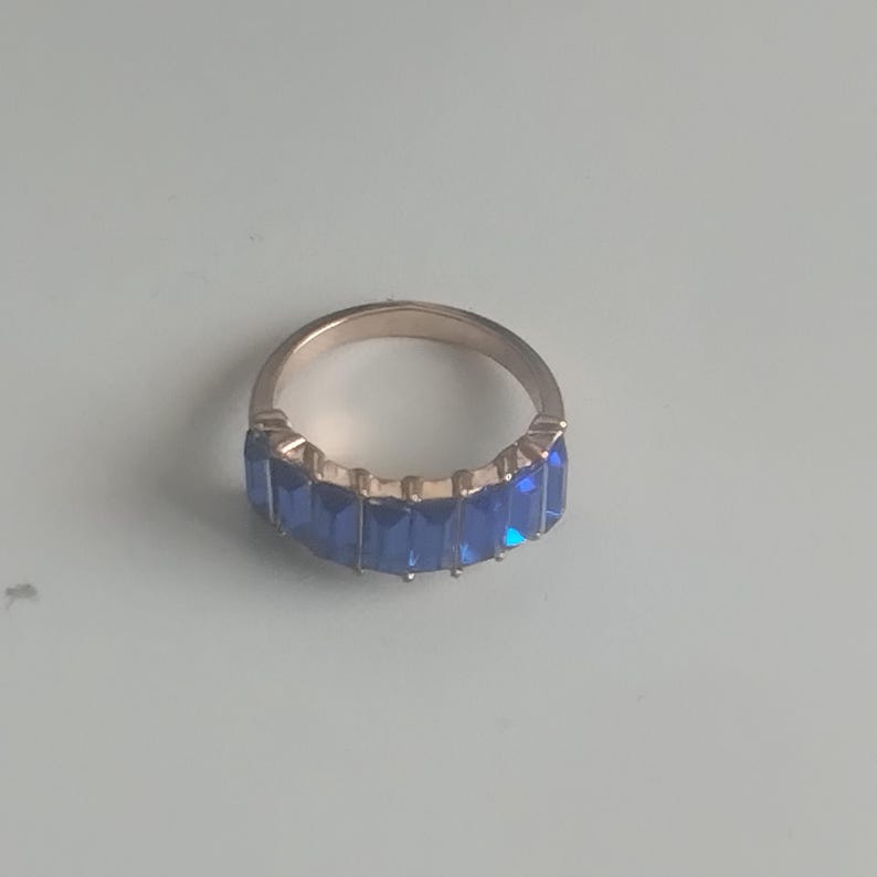 May include: A gold-toned ring featuring a row of rectangular, deep blue gemstones. The ring's band is a simple, polished metal, and the gemstones are set closely together, creating a continuous line of color. The ring is displayed on a light blue surface.