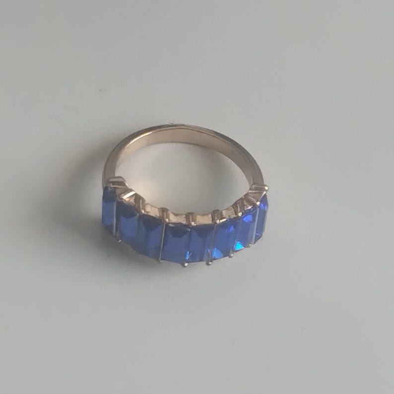 May include: A gold-toned ring featuring a row of rectangular, deep blue gemstones. The ring's band is a warm gold color, and the gemstones are set closely together, creating a continuous line of vibrant blue. The ring is a piece of jewelry.