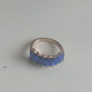 May include: A gold-toned ring featuring a band of rectangular, deep blue gemstones. The ring's design showcases a continuous row of vibrant blue stones, creating a striking contrast against the gold metal.