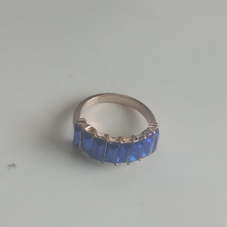 May include: A gold-toned ring featuring a band of rectangular, deep blue gemstones. The ring's design showcases a continuous row of vibrant blue stones, creating a striking contrast against the gold metal.