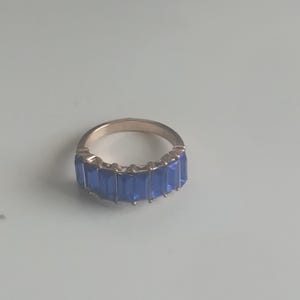 May include: A gold-toned ring featuring a row of rectangular, vibrant blue gemstones. The ring's band has a smooth, polished finish, and the blue stones are set closely together, creating a continuous line of color. The ring is displayed on a light-colored surface.