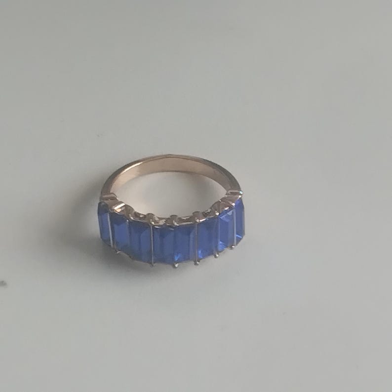 May include: A gold-toned ring featuring a row of rectangular, vibrant blue gemstones. The ring's band has a smooth, polished finish, and the blue stones are set closely together, creating a continuous line of color. The ring is displayed on a light-colored surface.