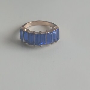 May include: A gold-toned ring featuring a band of rectangular, deep blue gemstones. The gemstones are set closely together, creating a continuous line of color around the ring's circumference. The ring is displayed on a light-colored surface.