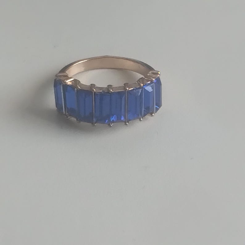 May include: A gold-toned ring featuring a band of rectangular, deep blue gemstones. The gemstones are set closely together, creating a continuous line of color around the ring's circumference. The ring is displayed on a light-colored surface.