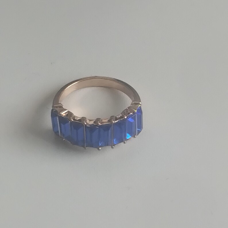 May include: A gold-toned ring featuring a row of rectangular, vibrant blue gemstones. The gemstones are set closely together, creating a continuous band of color. The ring's band has a smooth, polished finish.