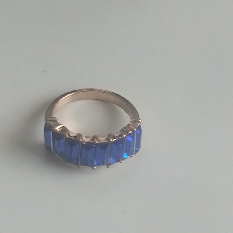 May include: A gold-toned ring featuring a row of rectangular, deep blue gemstones. The ring's band is a warm gold color, and the blue stones are set closely together, creating a continuous line of color. The ring is a piece of jewelry.