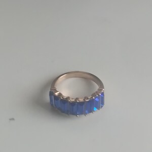 May include: A gold-toned ring featuring a row of rectangular, vibrant blue gemstones. The ring's band has a smooth, polished finish, and the gemstones are set closely together, creating a continuous line of color. This piece is a statement jewelry item.