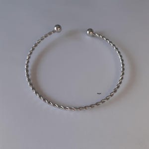 May include: A silver-coloured, open cuff bracelet. The bracelet features a twisted rope design and small, round ball ends. The jewellery is displayed on a white surface.