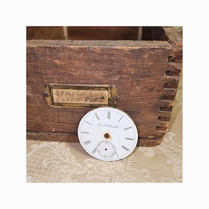 May include: A round white watch dial with black Roman numerals and a small sub-dial, positioned in front of a weathered wooden box with finger joints. A brass label holder on the box contains a paper insert with the handwritten text STAPLES COTER PINS.