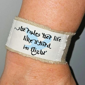 May include: A fabric bracelet with the text "She rules her life like a bird in flight" in black script. The bracelet has a white center with a light blue bird graphic and a beige, frayed edge.