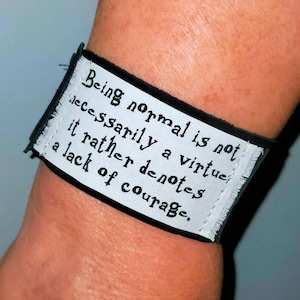 May include: A black and white wristband with the text "Being normal is not necessarily a virtue, it rather denotes a lack of courage." The wristband is wrapped around a wrist.
