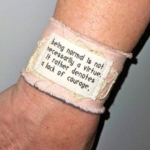 May include: A pink fabric wristband with the text "being normal is not necessarily a virtue, It rather denotes a lack of courage." The wristband has a lace trim and is wrapped around a wrist.