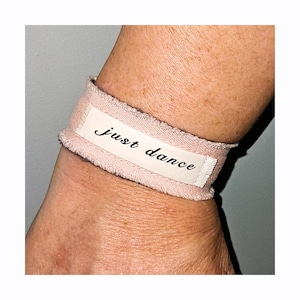 Puede incluir: Una pulsera de tela rosa claro con las palabras "just dance" impresas en una etiqueta blanca. La pulsera está envuelta alrededor de una muñeca.