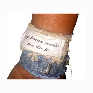 May include: A denim cuff bracelet featuring a frayed white fabric strip with the printed text "my boots made me do it". The accessory is accented with a small silver-toned metal cowboy boot charm attached to the blue denim fabric base.