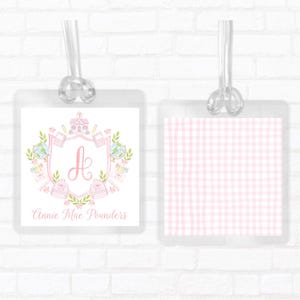 May include: Two square luggage tags with clear plastic loops. The left tag features a pink crest with the letter A, school-themed illustrations, and the name Annie Mae Pounders. The right tag displays a light pink and white gingham check pattern.