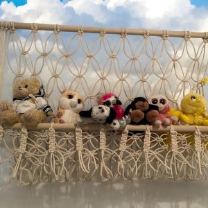 Könnte beinhalten: Ein weißer Makramee-Wandbehang mit sechs Stofftieren, die daran hängen. Die Tiere sind ein Bär, eine Katze, ein Flamingo, ein Affe, ein Hund und ein gelbes Küken.