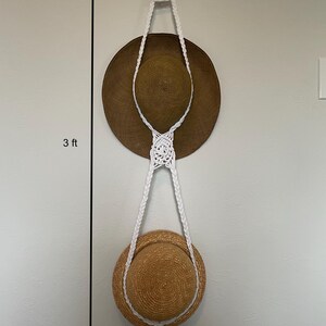 Puede incluir: Un perchero de macramé blanco colgado de una pared con dos sombreros de paja colgados. El perchero mide 91 cm de largo.