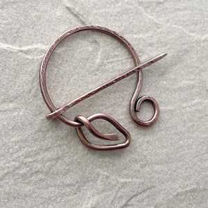May include: A copper-colored shawl pin with a hammered texture. The pin features a circular design with a straight pin and a decorative leaf-shaped element. The pin is designed to secure shawls or scarves.