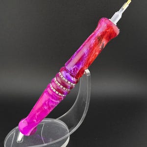 May include: A handcrafted pen with a vibrant design. The pen features a marbled effect in shades of red, pink, and purple, accented with a band of clear rhinestones. The pen is displayed on a clear acrylic stand against a black background.