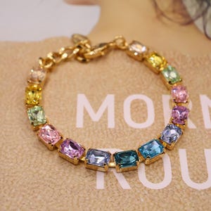 Op de afbeelding: Een goudkleurige armband met een reeks kleurrijke, rechthoekige edelstenen. De edelstenen zijn in een cirkelvormig patroon gerangschikt en tonen een spectrum aan kleuren, waaronder roze, blauw, groen en geel. De armband wordt gepresenteerd op een beige oppervlak.