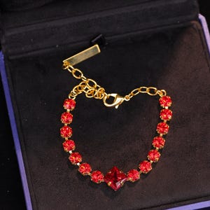 Pode incluir: Um bracelete de corrente dourada com cristais vermelhos quadrados. O bracelete está em uma caixa de veludo azul.