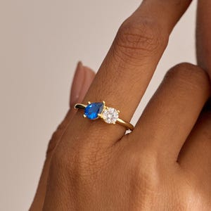Puede incluir: Un anillo de oro con una piedra preciosa azul y una piedra transparente de talla cuadrada. El anillo está engastado en una delgada banda de oro. Es una joya.