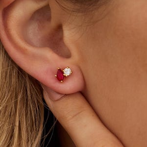 May include: Gold stud earrings with a red, teardrop-shaped gemstone and a round, clear gemstone. The earrings are small and delicate, designed to be worn in the ear. The earrings are set in gold.