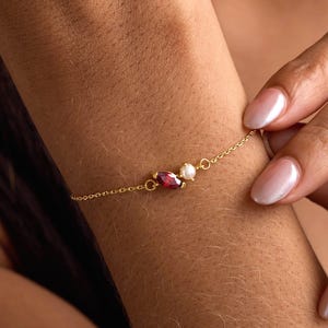 Könnte beinhalten: Ein zartes goldenes Armband mit einem kleinen, ovalen Rubin, einer weißen Perle und einem rosa Edelstein. Das Armband wird am Handgelenk getragen, eine Hand berührt es sanft. Das Armband ist durch eine feine Kette verbunden.