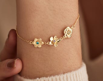 May include: A delicate gold bracelet featuring three flower charms. Each flower has a unique design, with one holding a blue gemstone, another a purple gemstone, and the last a clear gemstone. The bracelet is worn on a wrist.