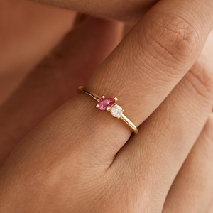 Può includere: Un delicato anello d'oro con una pietra preziosa rosa e una bianca. L'anello è indossato su un dito.