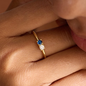 Op de afbeelding: Een gouden ring met een blauwe en een witte edelsteen. De ring heeft een dunne gouden band. De edelstenen zijn rond en dicht bij elkaar geplaatst. De ring zit om de vinger van een persoon.