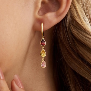 Könnte beinhalten: Goldene Creolen mit drei tropfenförmigen Edelsteinen in Rot, Gelb und Rosa. Die Ohrringe hängen und werden an einem Ohr getragen. Die Edelsteine sind in Gold gefasst und haben eine polierte Oberfläche.