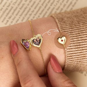 Könnte beinhalten: Ein goldenes Kettenarmband mit einem herzförmigen Medaillon, das sich öffnen lässt und zwei kleine Fotos im Inneren zeigt. Ein kreisförmiger Ausschnitt zeigt das geschlossene Medaillon mit einem kleinen roten Stein in der Mitte.